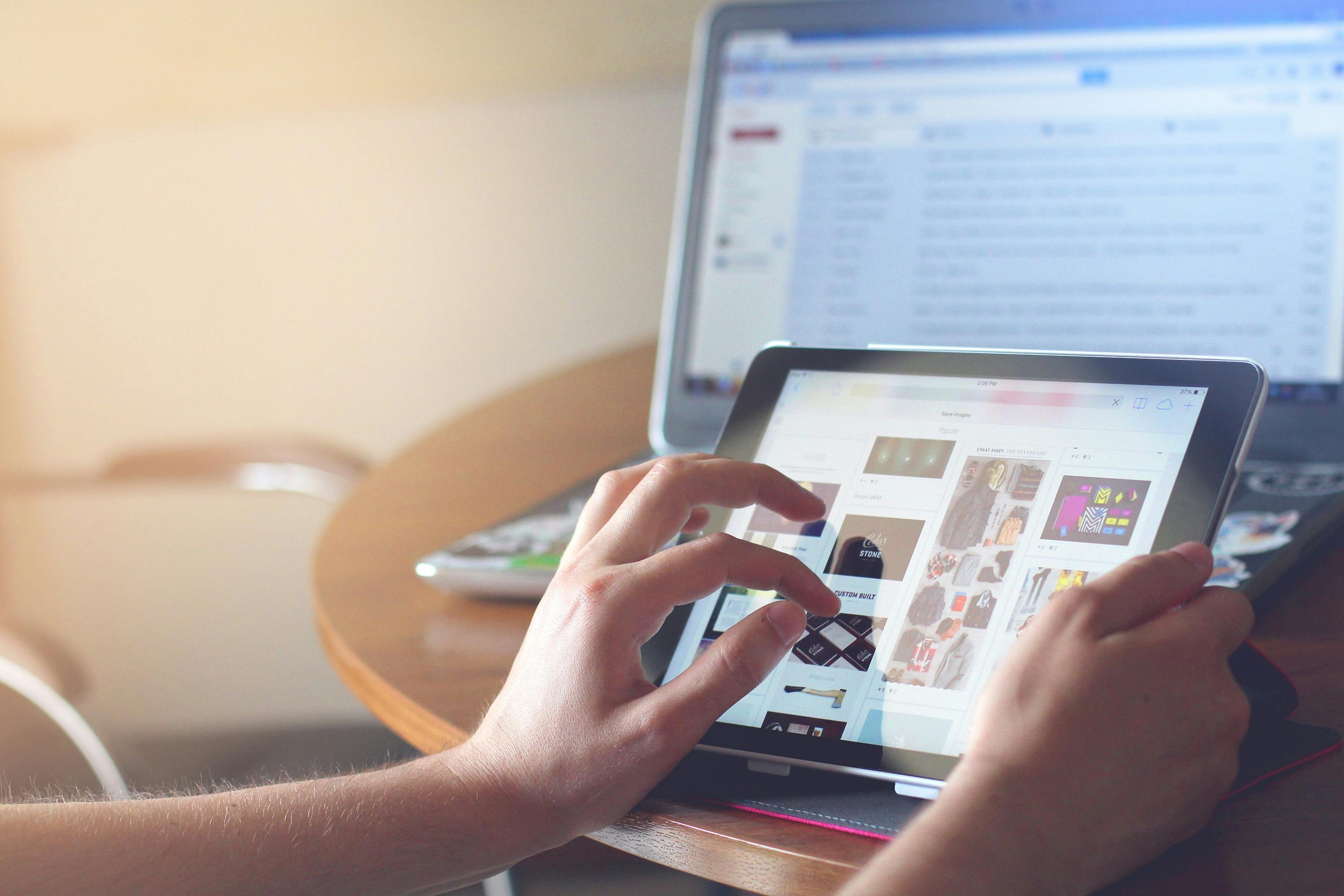 A close-up of a person's hands using a tablet to scroll through a gallery of images, with an open laptop displaying text in the background.